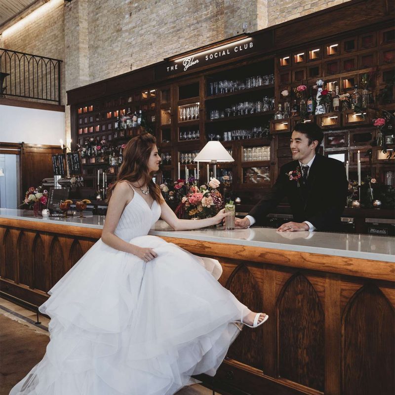 Bride sits at the bar while groom serves her a drink.