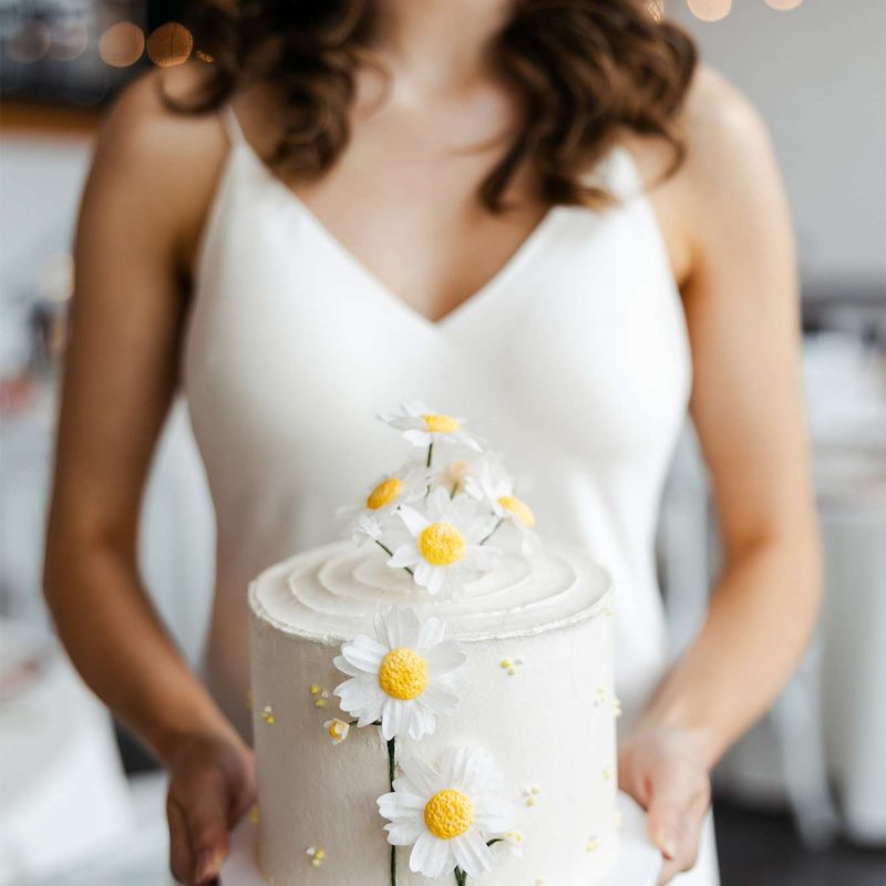 Bride holds vegan wedding cake with white buttercream and daisy flowers (crafted from edible wafer paper)
