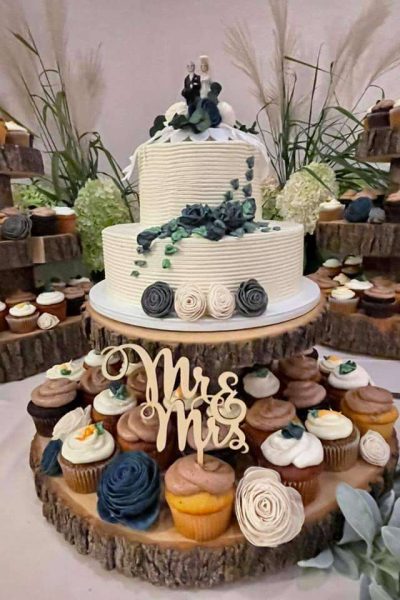 Two tiered wedding cake with topper over rustic display of cupcakes with floral accents