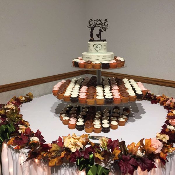 Sweets table with three levels of cupcakes displayed under two tier wedding cake with wedding topper and fall color floral arrangement on table edge