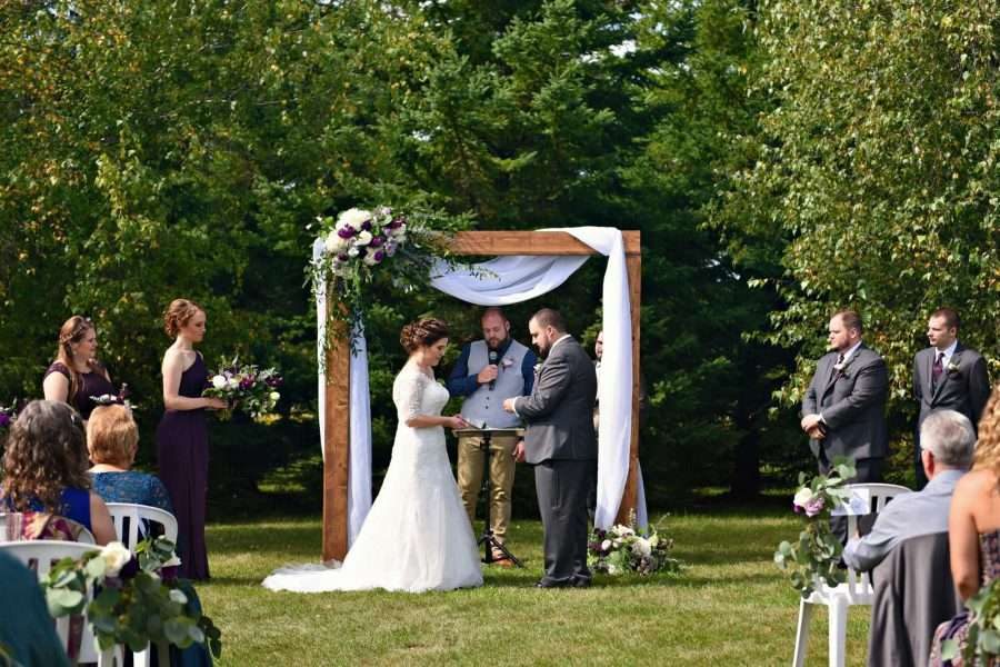 Couple exchange vows under wooden arch adorned with lush florals and white drapery.