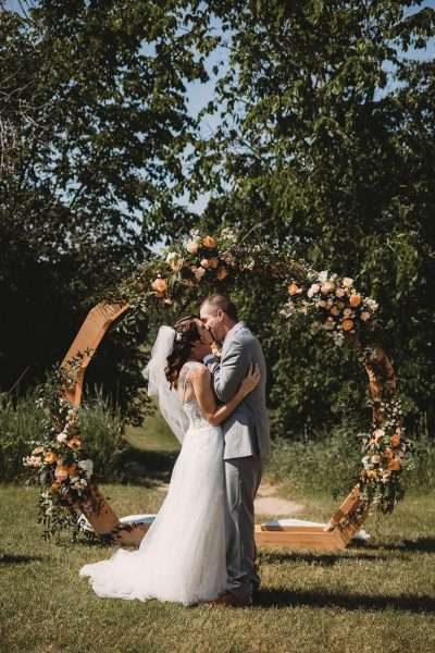 Bride and groom kiss under curved floral drenched arch at outdoor wedding ceremony