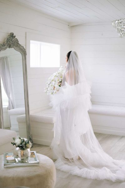 Veiled bride looks into full length mirror.
